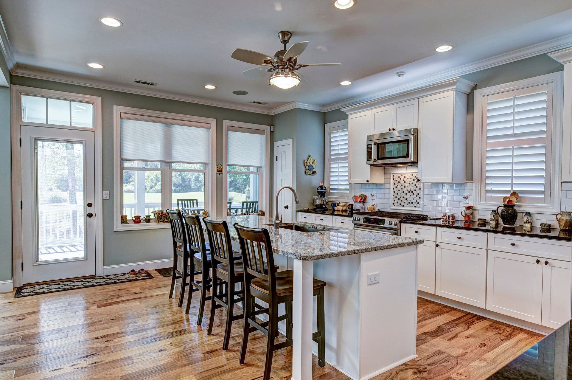 A modern kitchen with white cabinets, granite island, and wooden floor. Bar stools line the island, and large windows brighten the space.