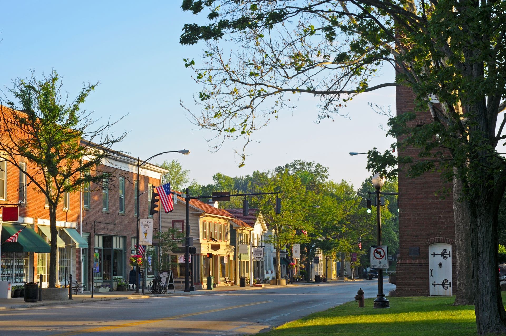 A quaint, sunlit small-town street with colorful brick buildings, trees lining the road, and American flags. The scene is peaceful and inviting.