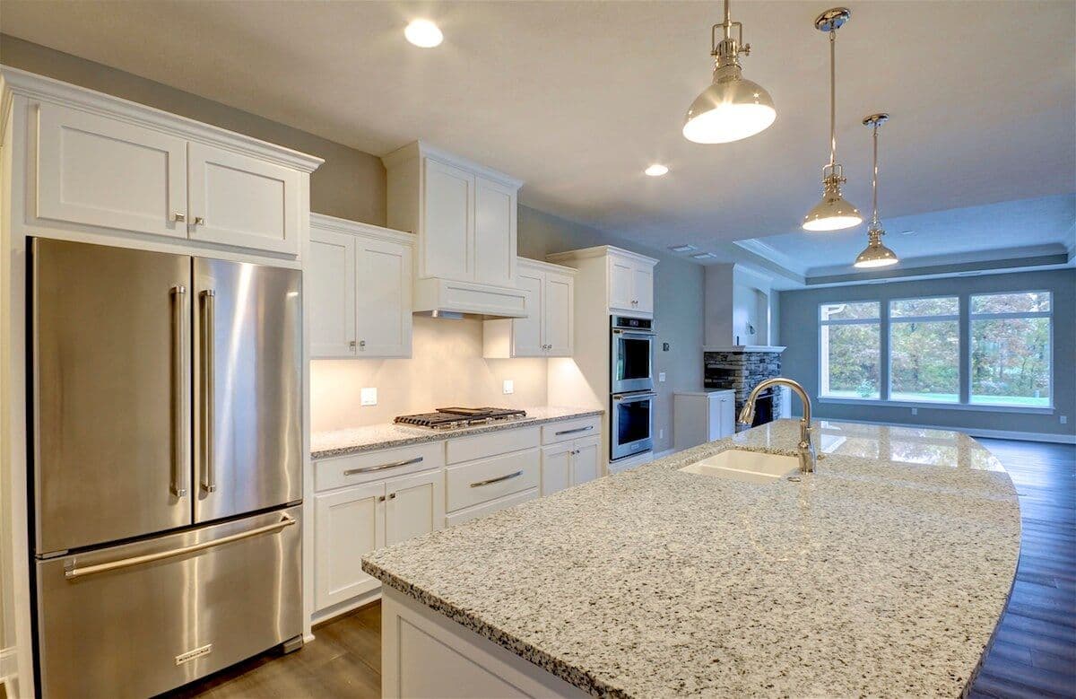 Spacious kitchen with white cabinets, stainless steel appliances, large granite island, and pendant lights. Sunlight streams through big windows.