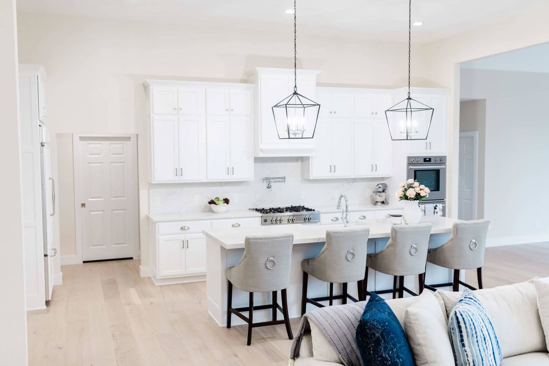 Spacious, modern kitchen with white cabinetry, stainless steel appliances, and a large island with four gray chairs. Two pendant lights hang above.