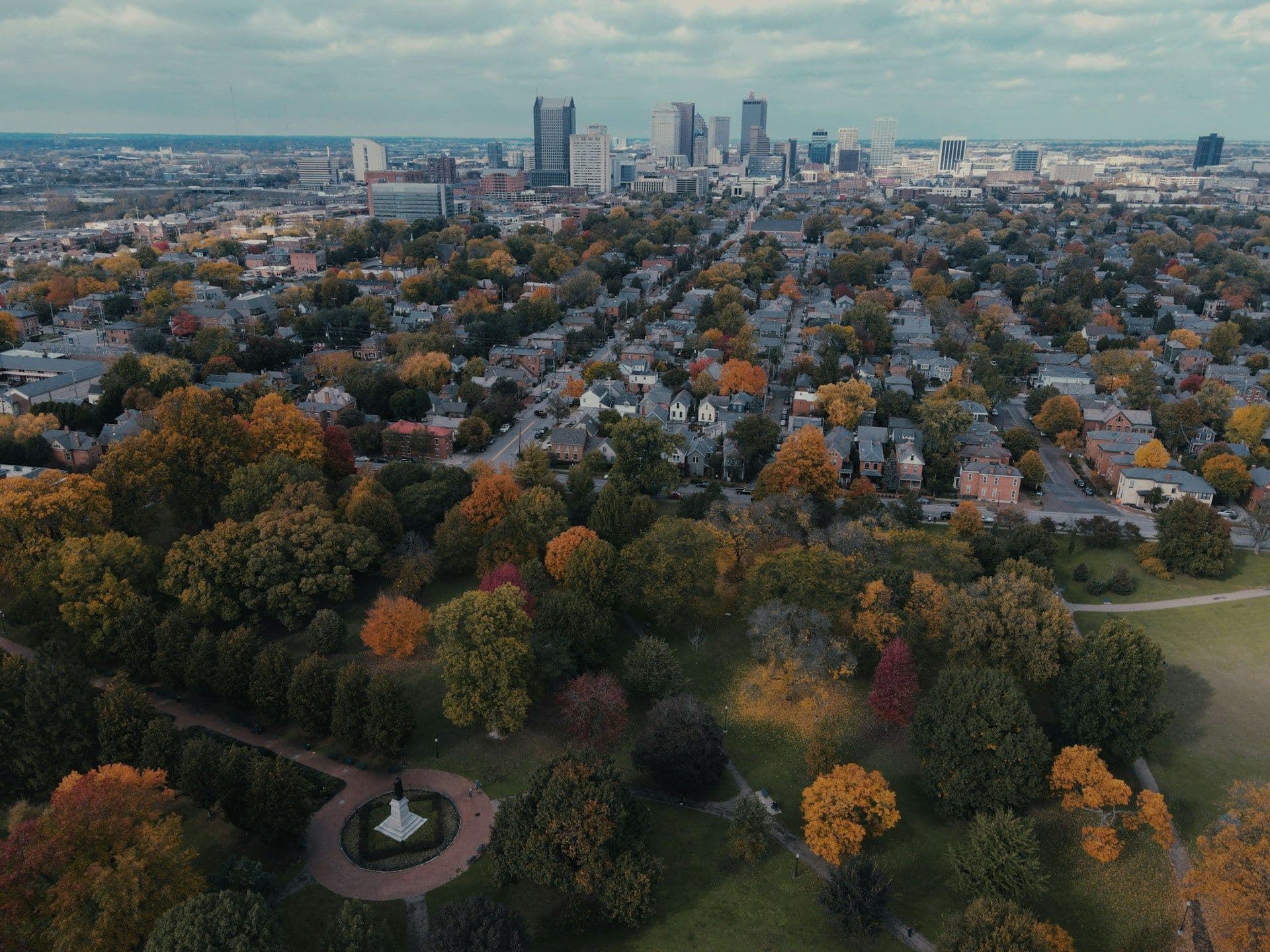 Aerial view of an Ohio cityscape with a park in the foreground. Autumn trees with colorful leaves contrast the dense residential area and distant skyscrapers.